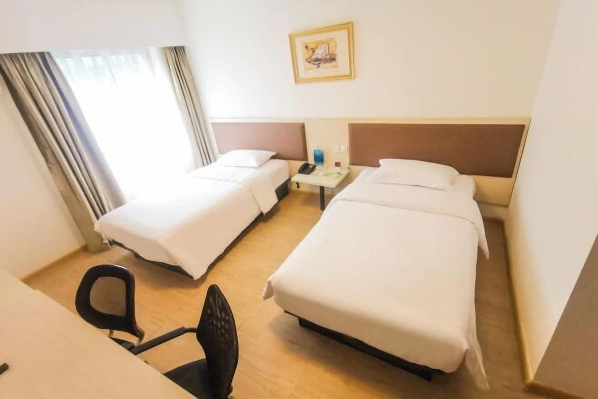 A hotel room with two single beds, a desk with a chair, a small table with a phone and a bottle, and a window with curtains.