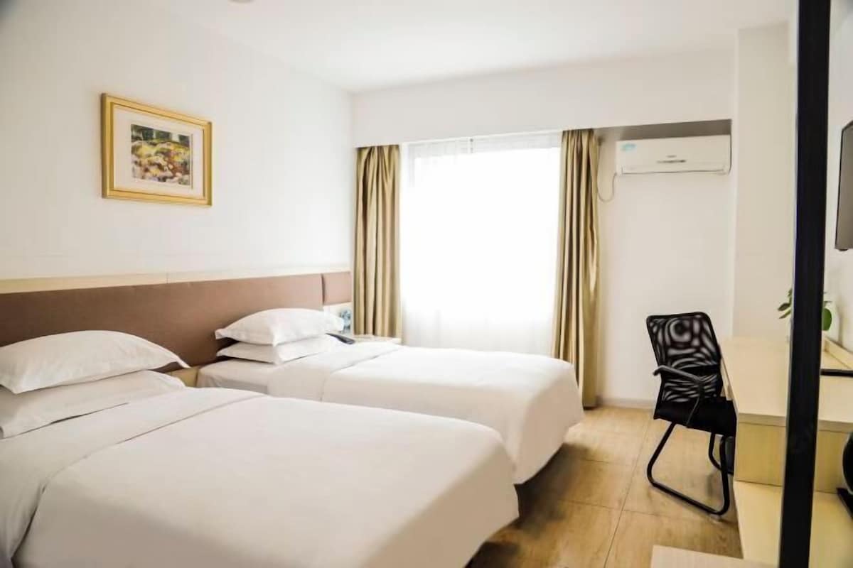 A hotel room with two beds, a desk with a chair, a window with curtains, and a framed picture on the wall.