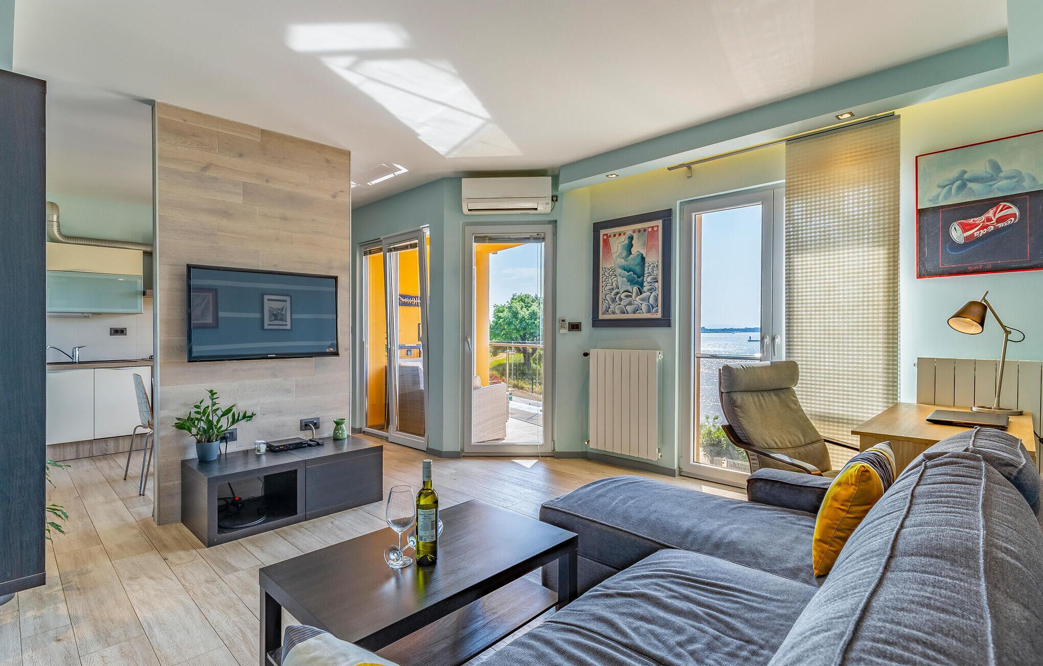 Sala de estar moderna com sofá, mesa de centro e TV na parede. Há uma porta de vidro deslizante que dá para uma varanda com vista para o mar.