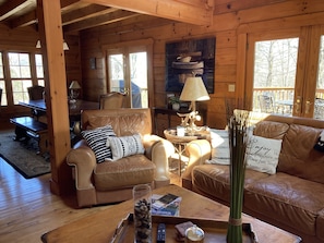A cozy cabin interior with a wooden interior, leather sofas, a dining table, and a view of the outdoors through large windows.