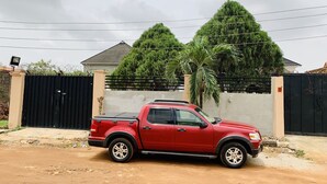 A red pickup truck parked in front of a gated property with a house and lush greenery in the background. - Elegantly Apportioned 4-bed House in Ikorodu (Ikorodu)