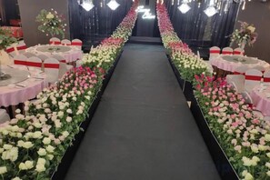 Una larga mesa con un camino de mesa negro, rodeada de sillas blancas y rosas, y un arreglo floral a cada lado.