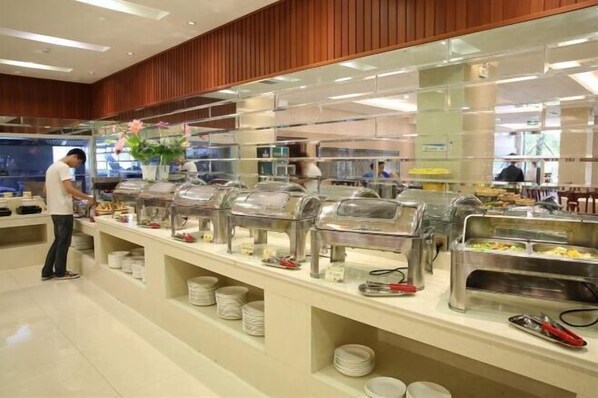 Zona de bufé con varias estaciones de autoservicio, cada una con recipientes cubiertos para la comida. Hay platos y cubiertos en la barra, y se ve a una persona preparando alimentos.