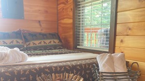 A wooden cabin room with a bed, pillows, a small table with a basket, and a window with blinds. - Bearadise in the Red, Wifi, Hot Tub, great proximity to Red River Gorge (Campton)