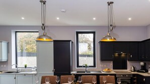 A modern kitchen with a dining area, featuring a large wooden table, six chairs, pendant lighting, and a view of the outdoors through large windows. - BOOTique House - Luxury Group Accommodation for 12 (Wakefield)