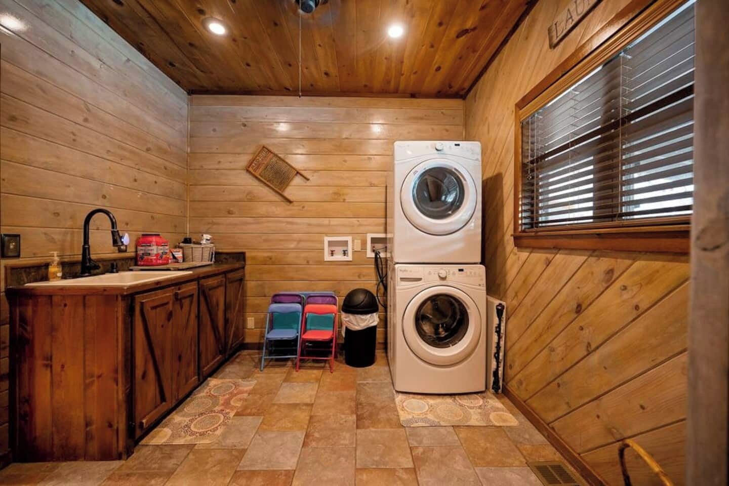 A compact, wooden-floored laundry room with stacked washers and dryers, a sink, and a window with blinds.