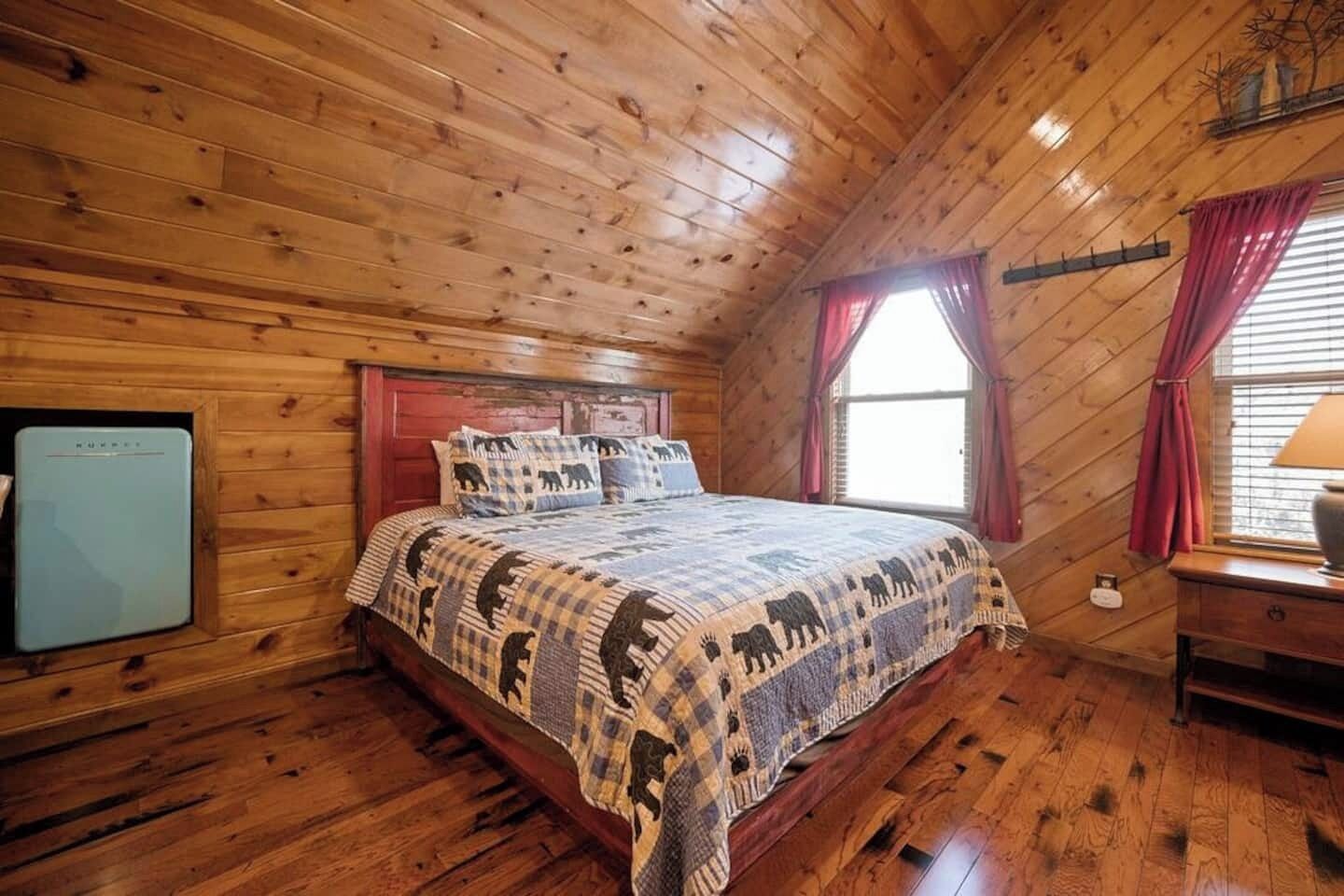 Un dormitorio de cabaña de madera con una cama, una mesita y un ventanal con cortinas.