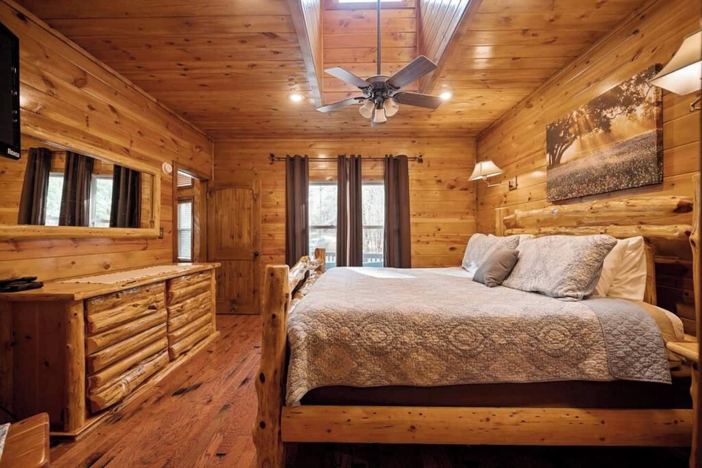Ein Zimmer in einer Holzhütte mit einem Bett, einem Schminktisch, einem Deckenventilator und einem Fenster mit Vorhängen.