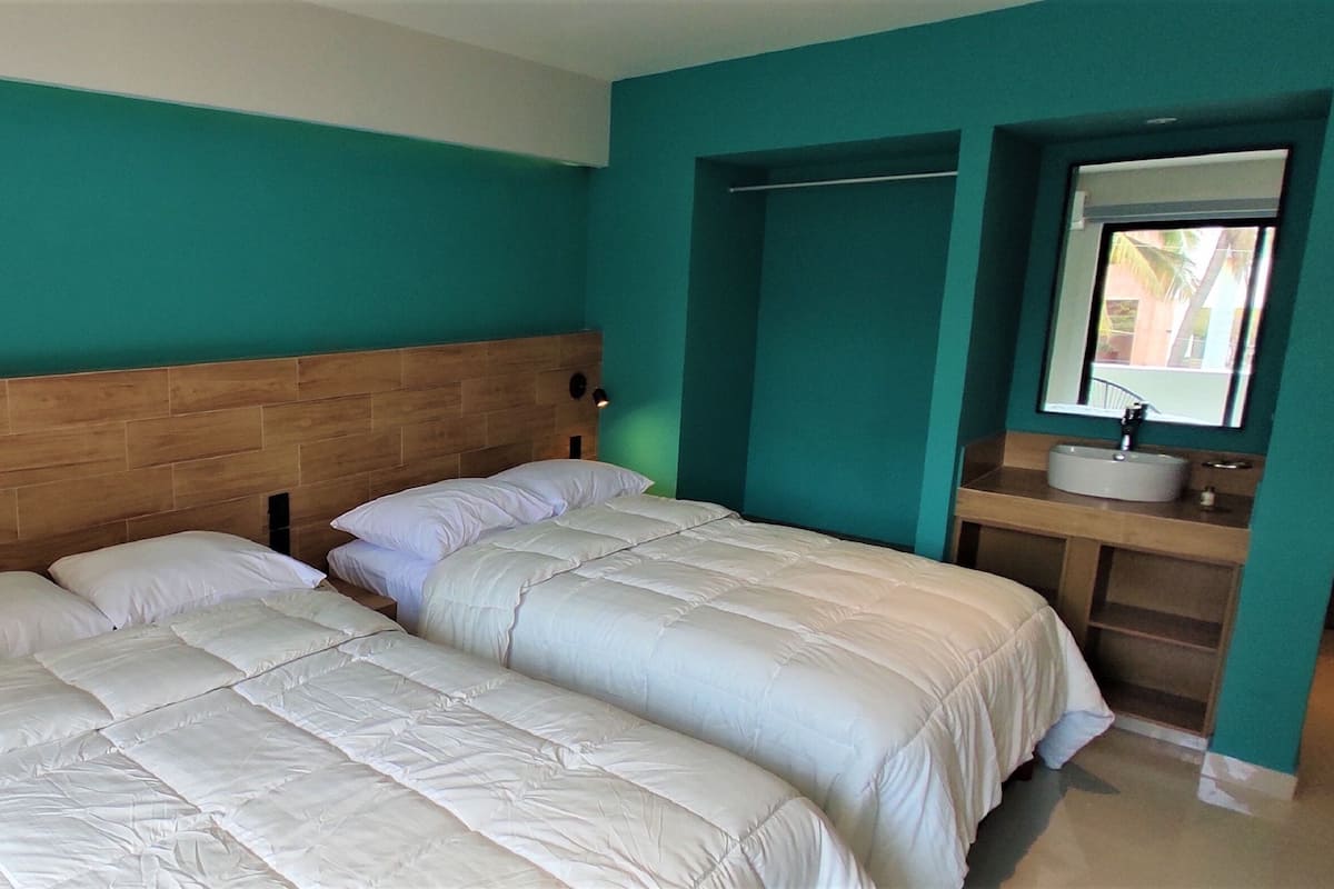 A room with two beds, a wooden headboard, a teal-colored wall, a bathroom with a sink and mirror, and a small window.