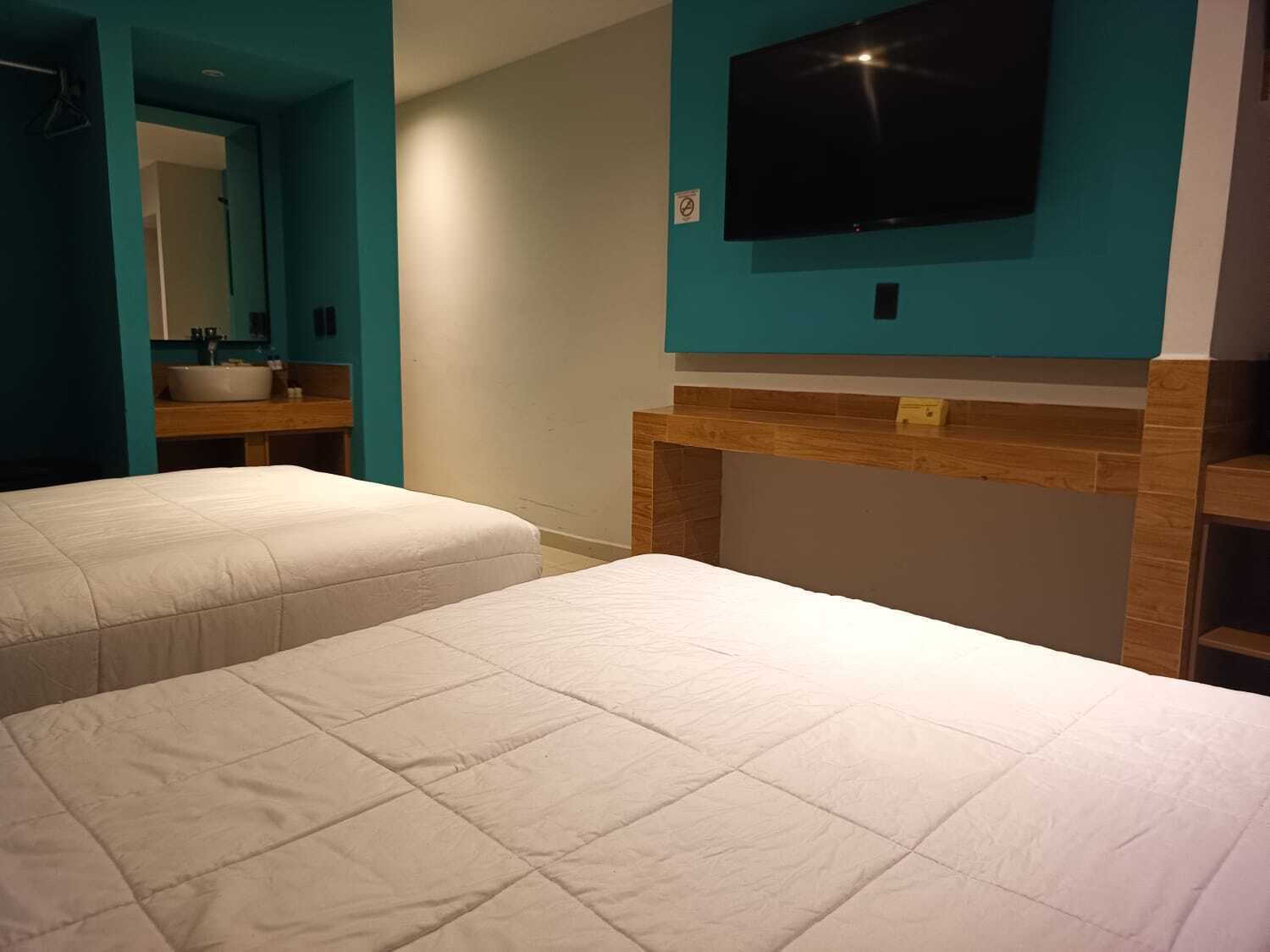 A hotel room with two beds, a TV mounted on the wall, and a bathroom with a sink and mirror.