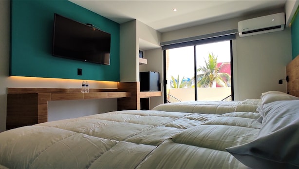 Ein Hotelzimmer mit einem großen Bett, einem flachbildfernseher an der Wand, einer Minibar und einem großen Fenster mit Blick auf Palmen.