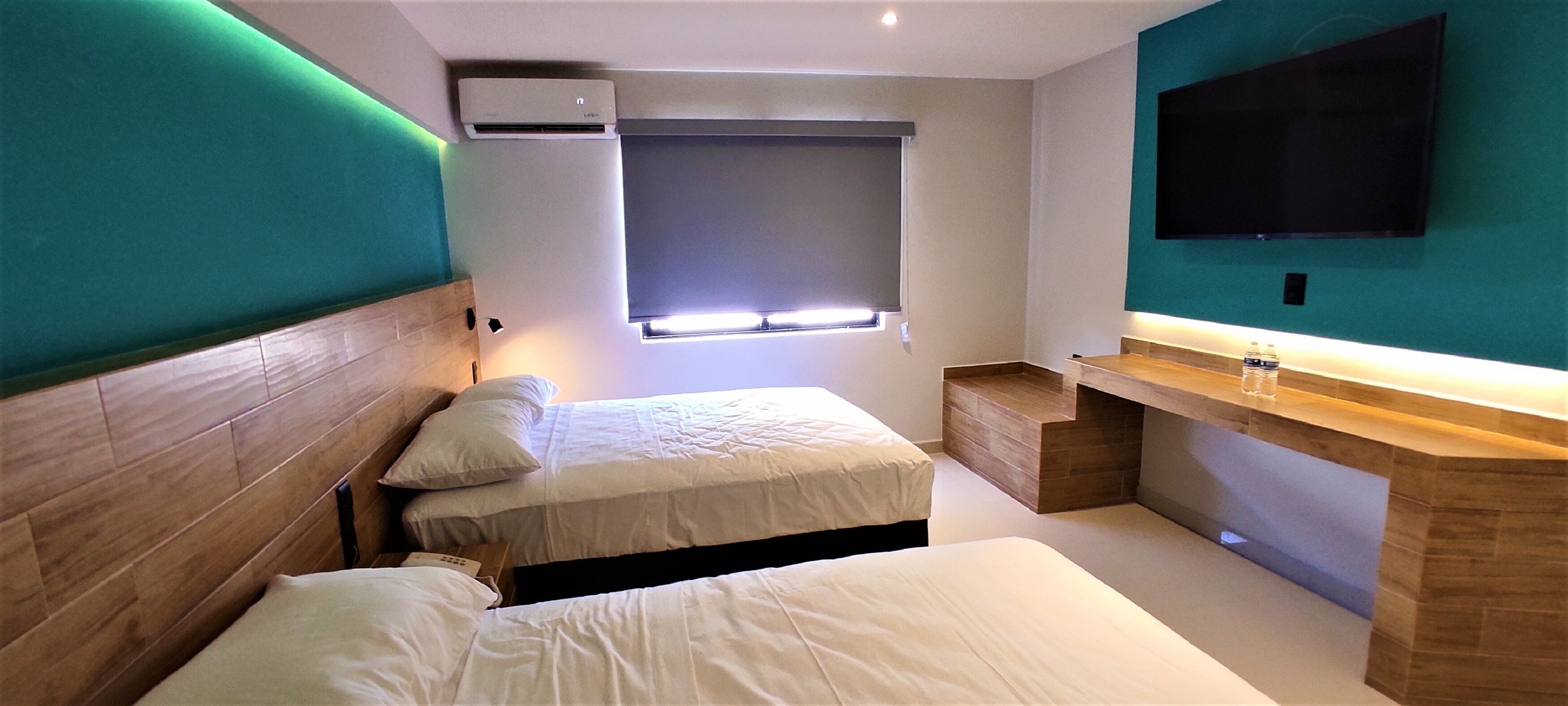 A modern hotel room with two beds, a built-in desk, a TV, and a window with blinds.