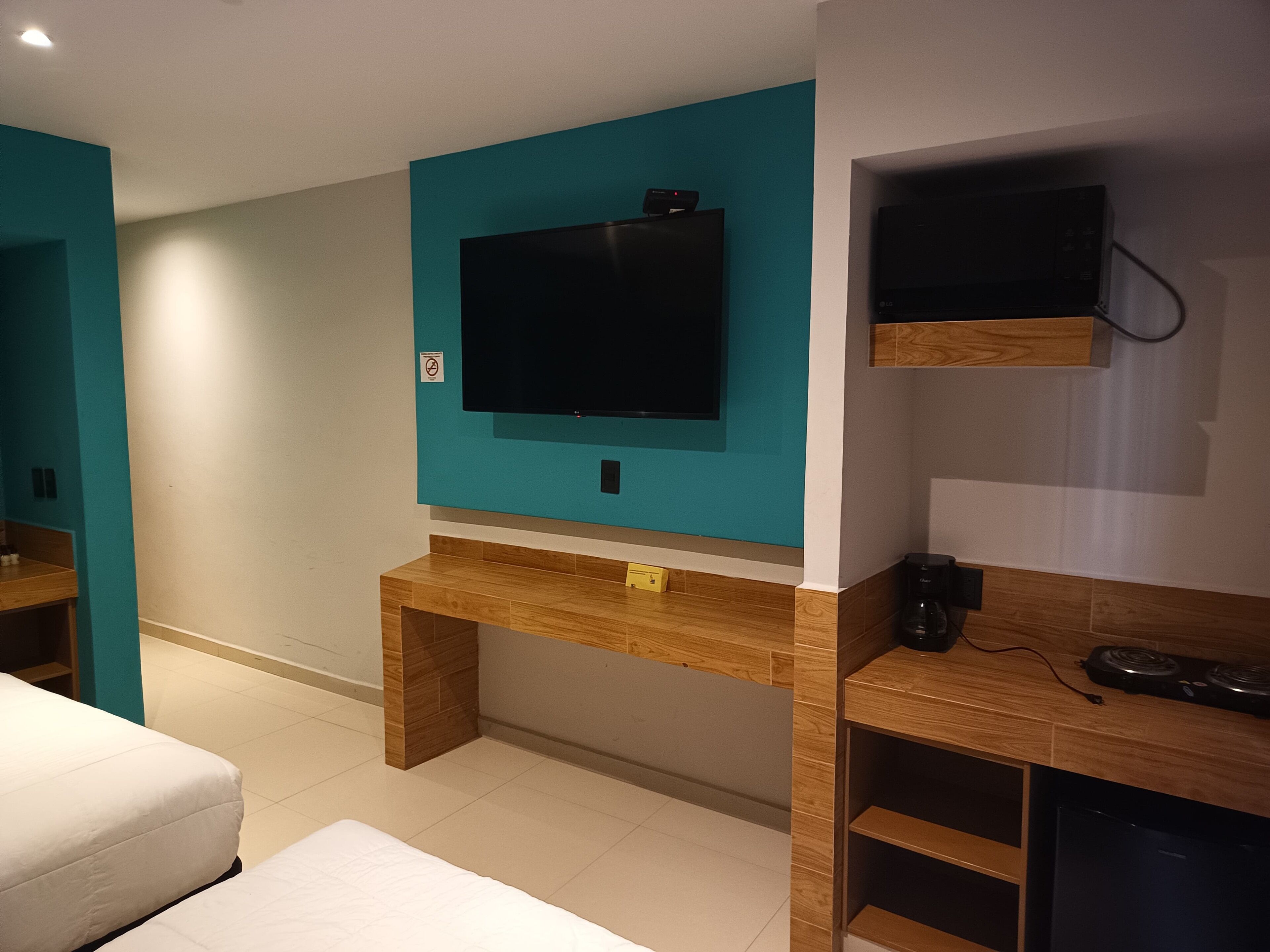 A modern hotel room with a teal accent wall, a large flat-screen TV mounted on a wooden console, a small coffee machine, and a built-in mini-fridge.