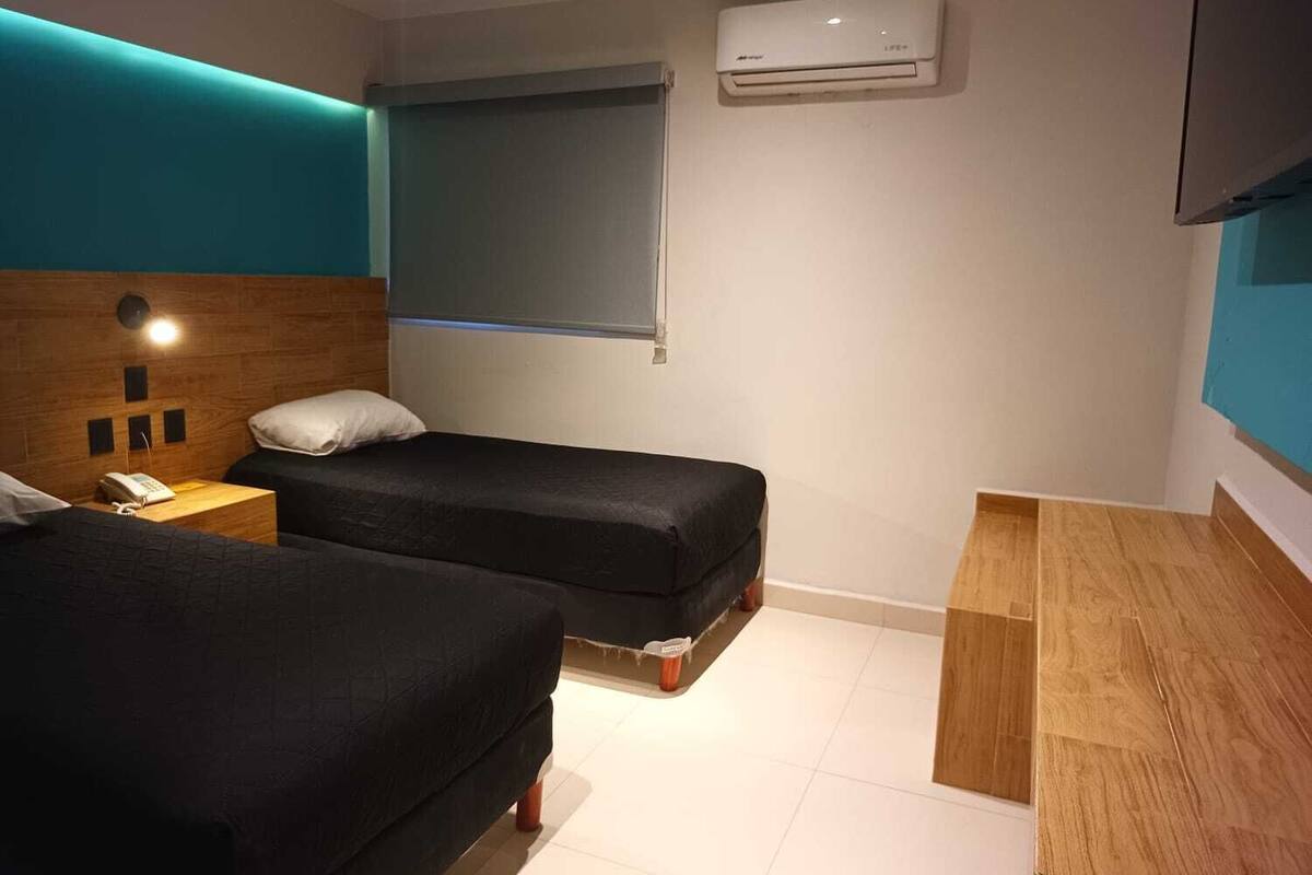 A hotel room with two beds, a wooden headboard, a bedside table with a lamp, an air conditioner, and a TV mounted on the wall.