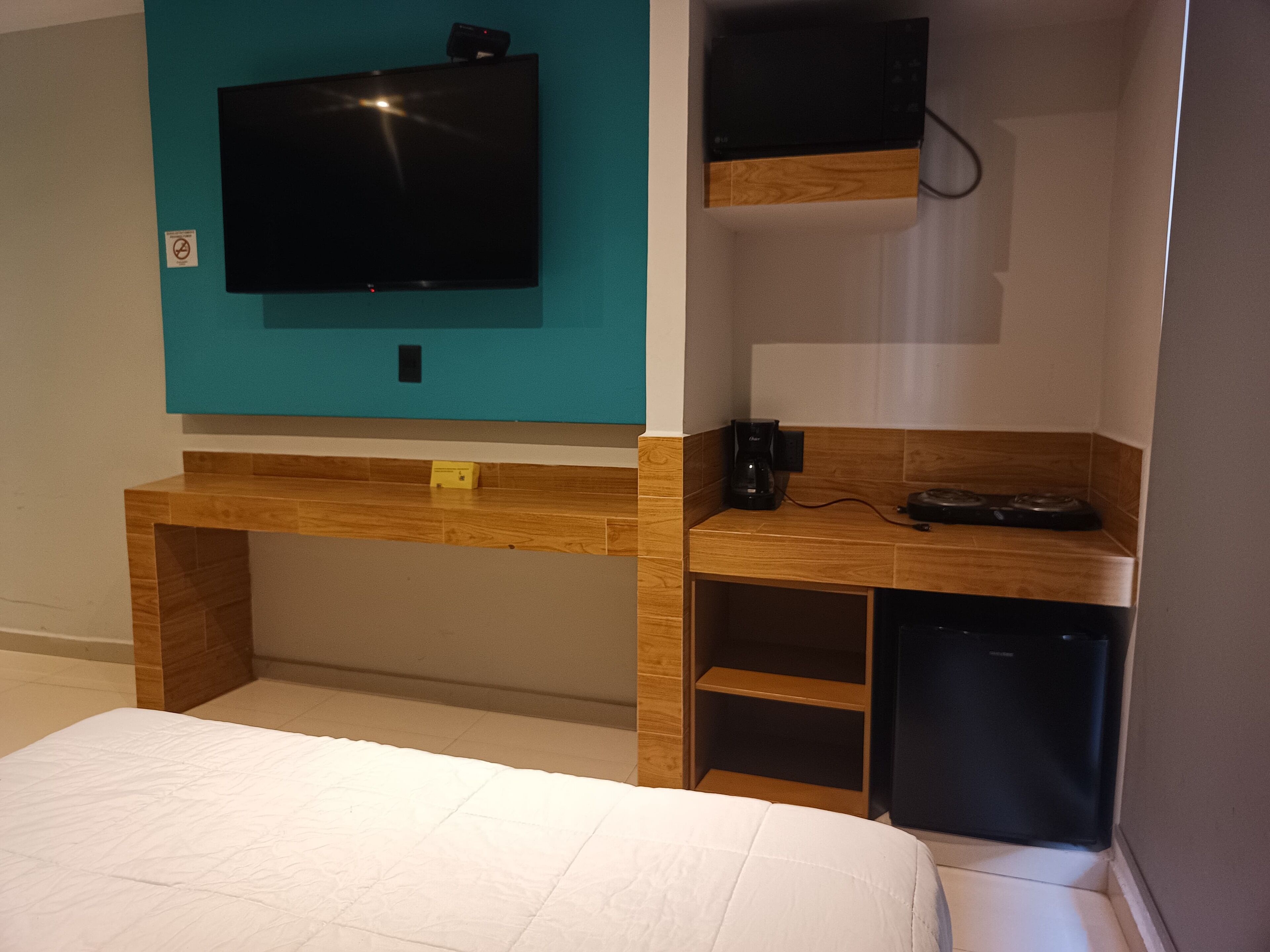 Une chambre à coucher avec un lit, un téléviseur fixé au mur, qui est de couleur turquoise, un bureau en bois avec une machine à café et un petit appareil électroménager noir.