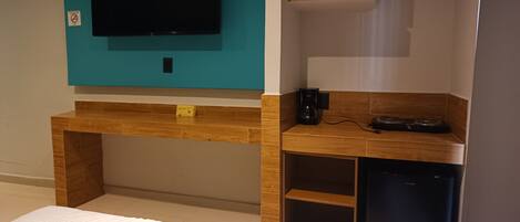 Une chambre à coucher avec un lit, un téléviseur fixé au mur, qui est de couleur turquoise, un bureau en bois avec une machine à café et un petit appareil électroménager noir.