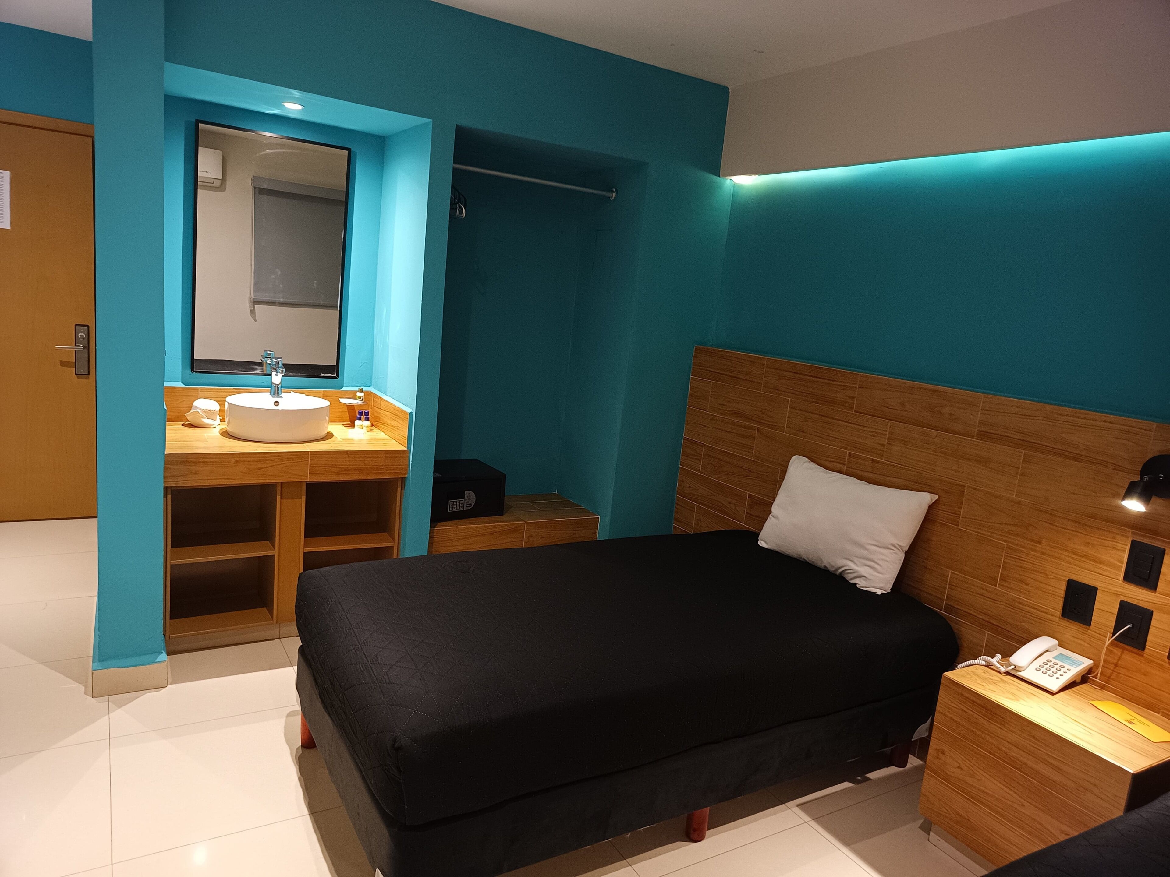 A modern bedroom with a wooden headboard, a black bedspread, a white pillow, a bedside table with a phone, and a bathroom with a sink and mirror.