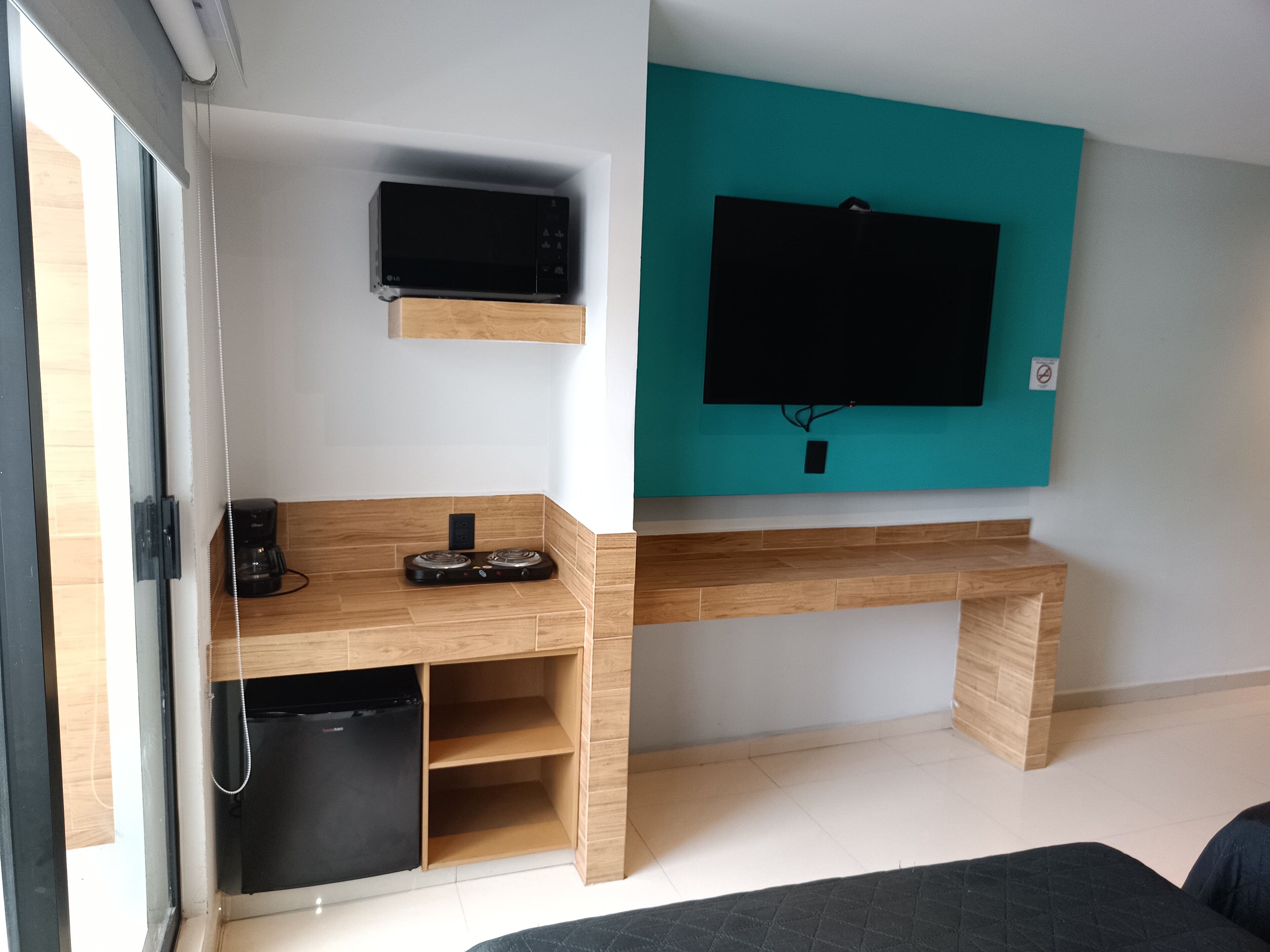 A modern living room with a flat-screen TV mounted on a teal wall, a wooden desk with a coffee maker, and a black refrigerator.