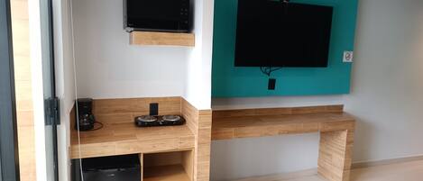A modern living room with a flat-screen TV mounted on a teal wall, a wooden desk with a coffee maker, and a black refrigerator.