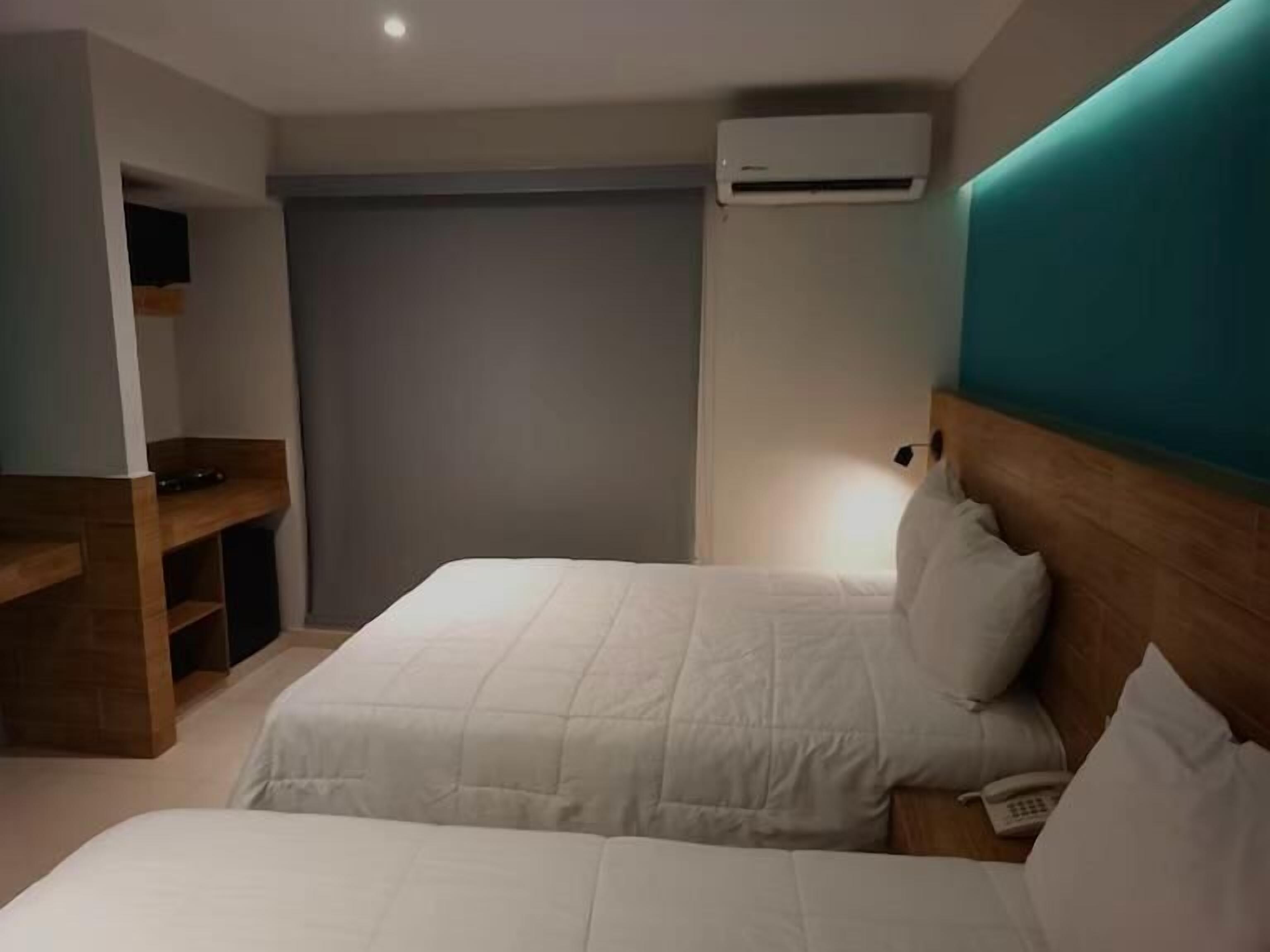 A hotel room with two beds, a wooden desk, a wardrobe, and an air conditioning unit.