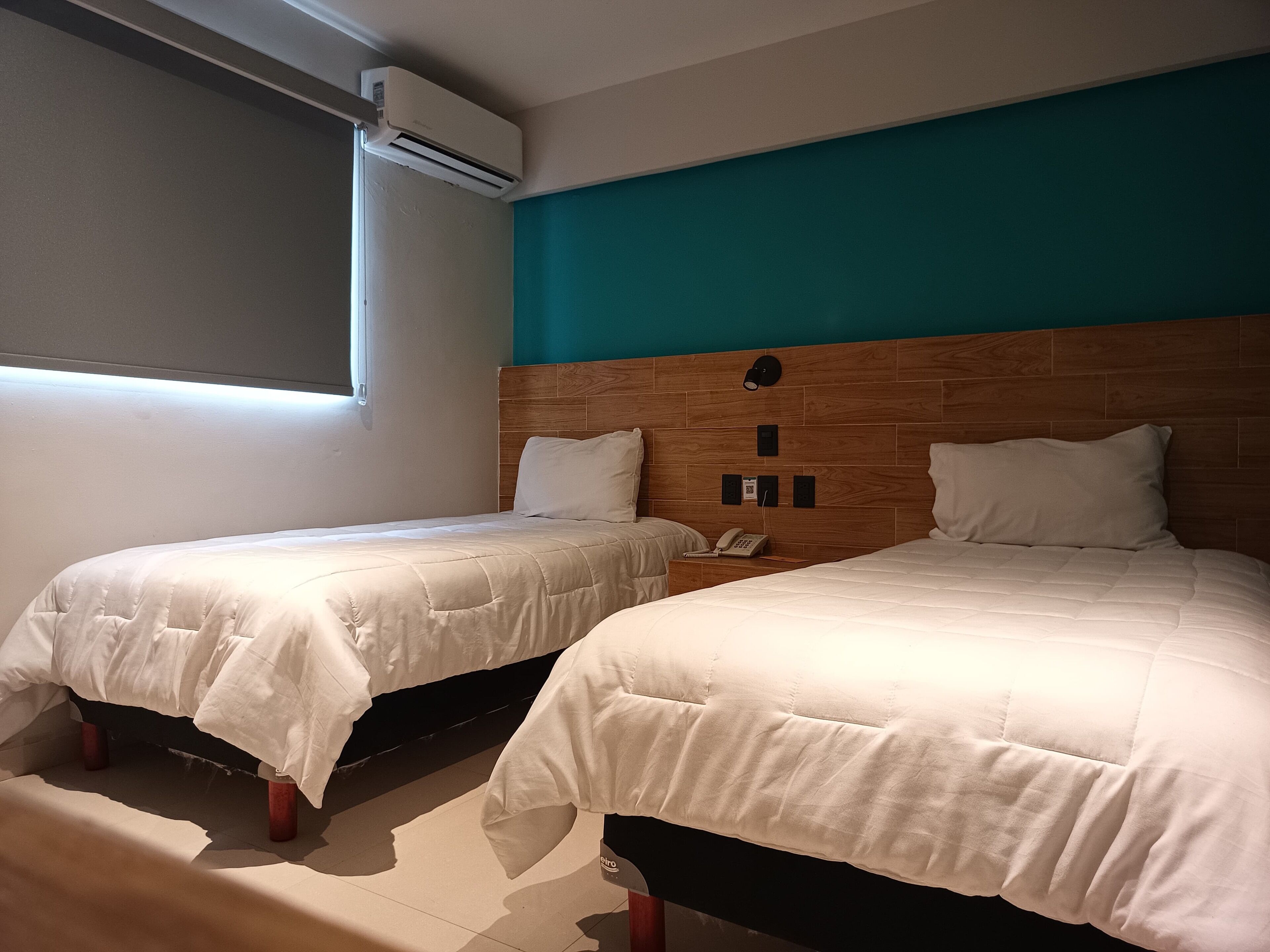 Deux lits simples avec des têtes de lit en bois, un climatiseur fixé au mur et un téléphone sur la table de chevet.