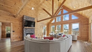 Un salón amplio con un sofá seccional en forma de L, una chimenea de piedra y grandes ventanales que ofrecen vistas al exterior.