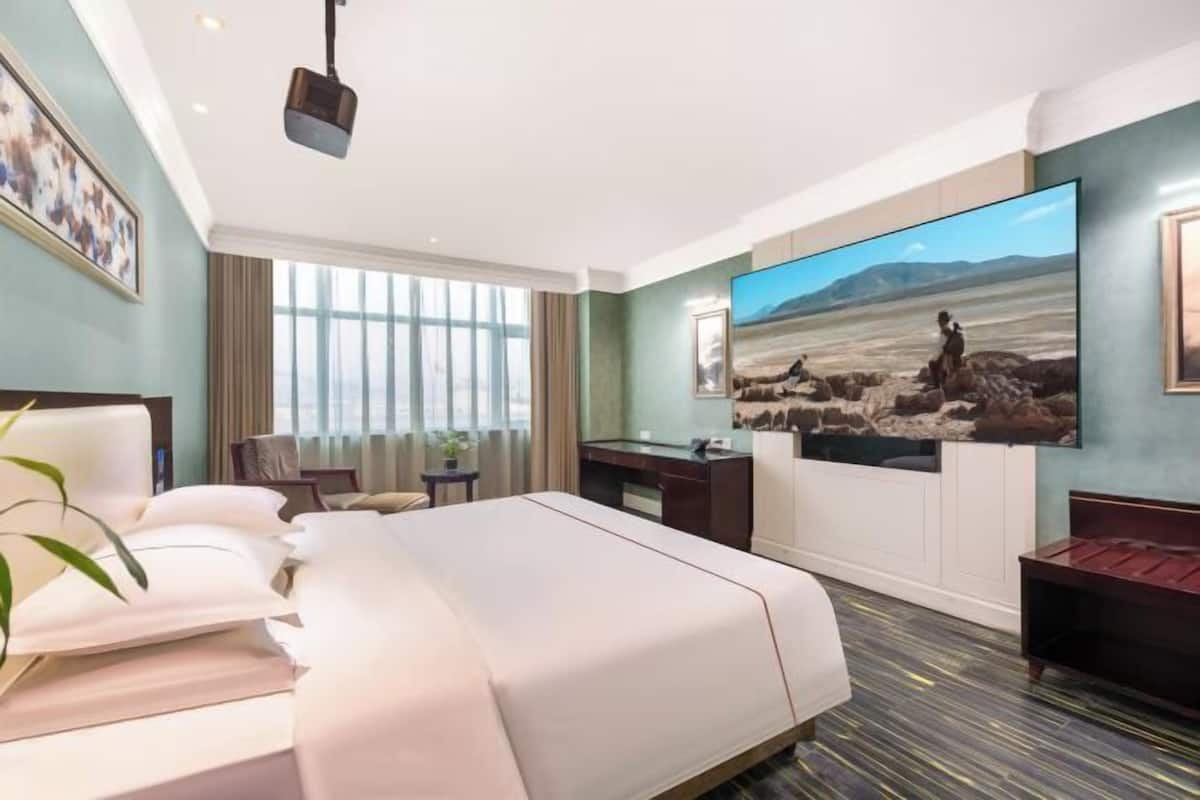 A hotel room with a large bed, a desk, a chair, a TV, and a window with curtains.