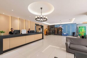 Zona de recepción de un hotel con un mostrador, dos recepcionistas, una araña y un área de descanso con un sofá.