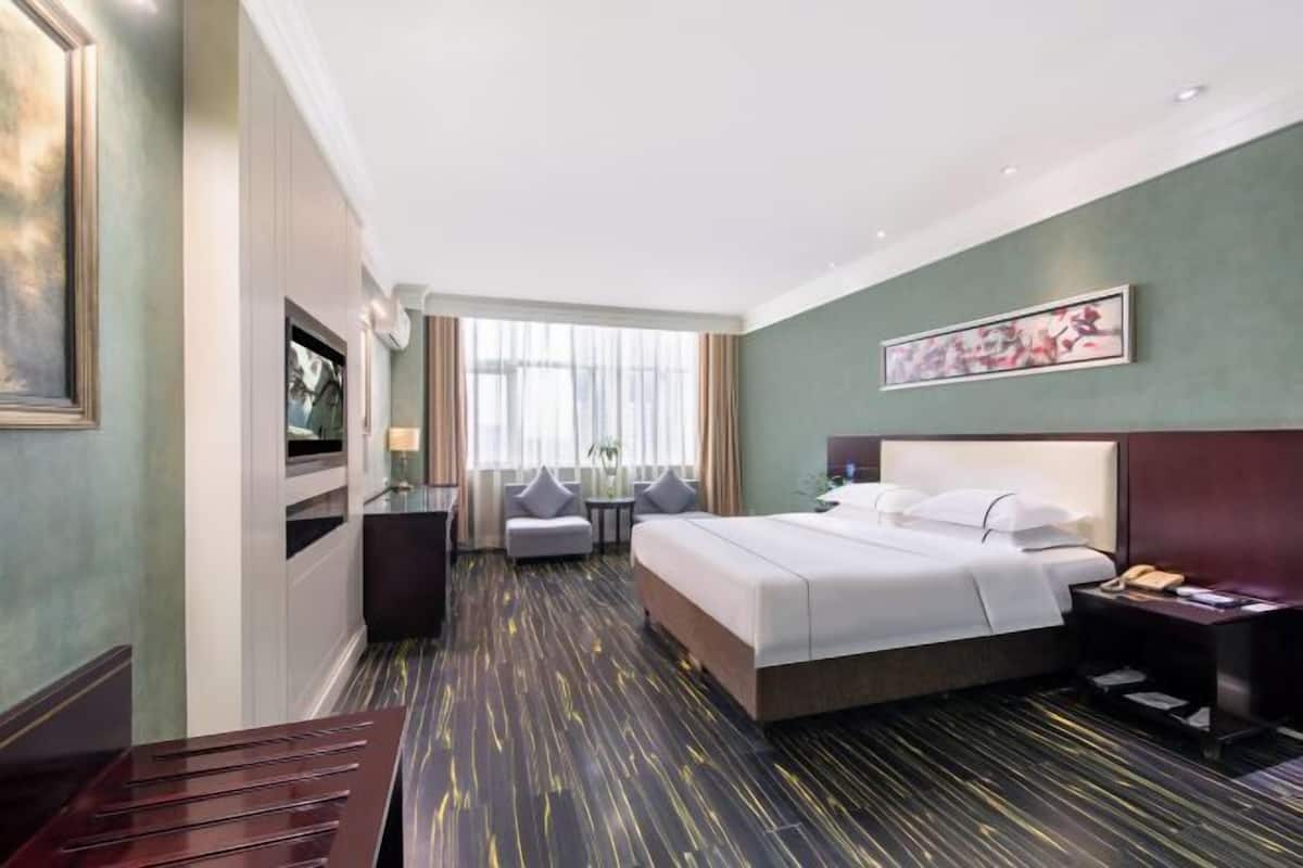 A hotel room with a bed, bedside table, lamp, and a view of the city through large windows.