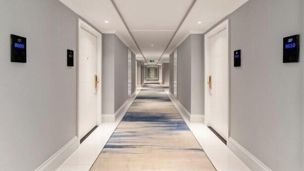 En lang, smal hotelgang med døre på begge sider, blåt tæppe og digitale nummerplader til værelserne.