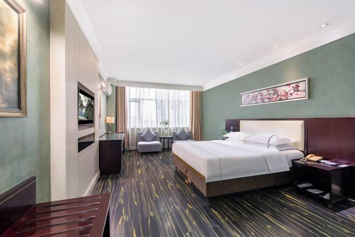 A hotel room with a large bed, a desk, a chair, and a view of the city through large windows.