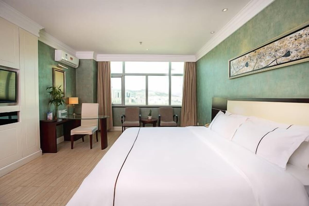 A hotel room with a large bed, a desk with a chair, a TV, and a window with a city view.