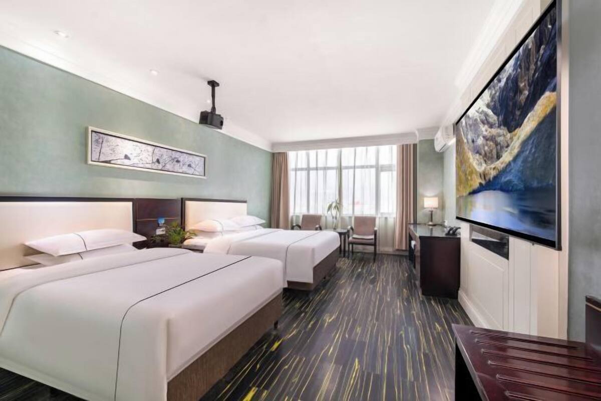 A hotel room with two beds, a TV, a painting, and a window.