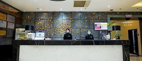 A hotel reception area with a marble counter, two receptionists, a digital display, and a wall decorated with autumn leaves.