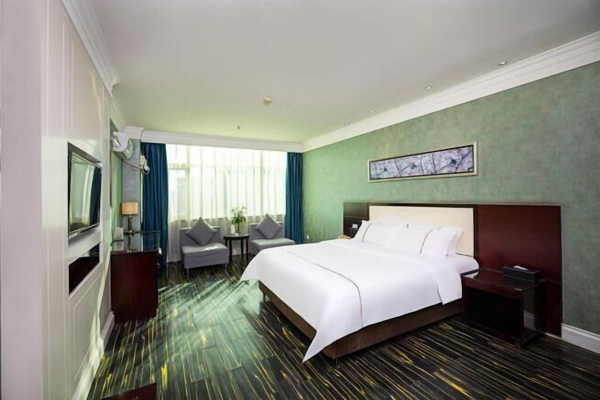 A hotel room with a large bed, a TV, a desk, a chair, and a window with curtains.
