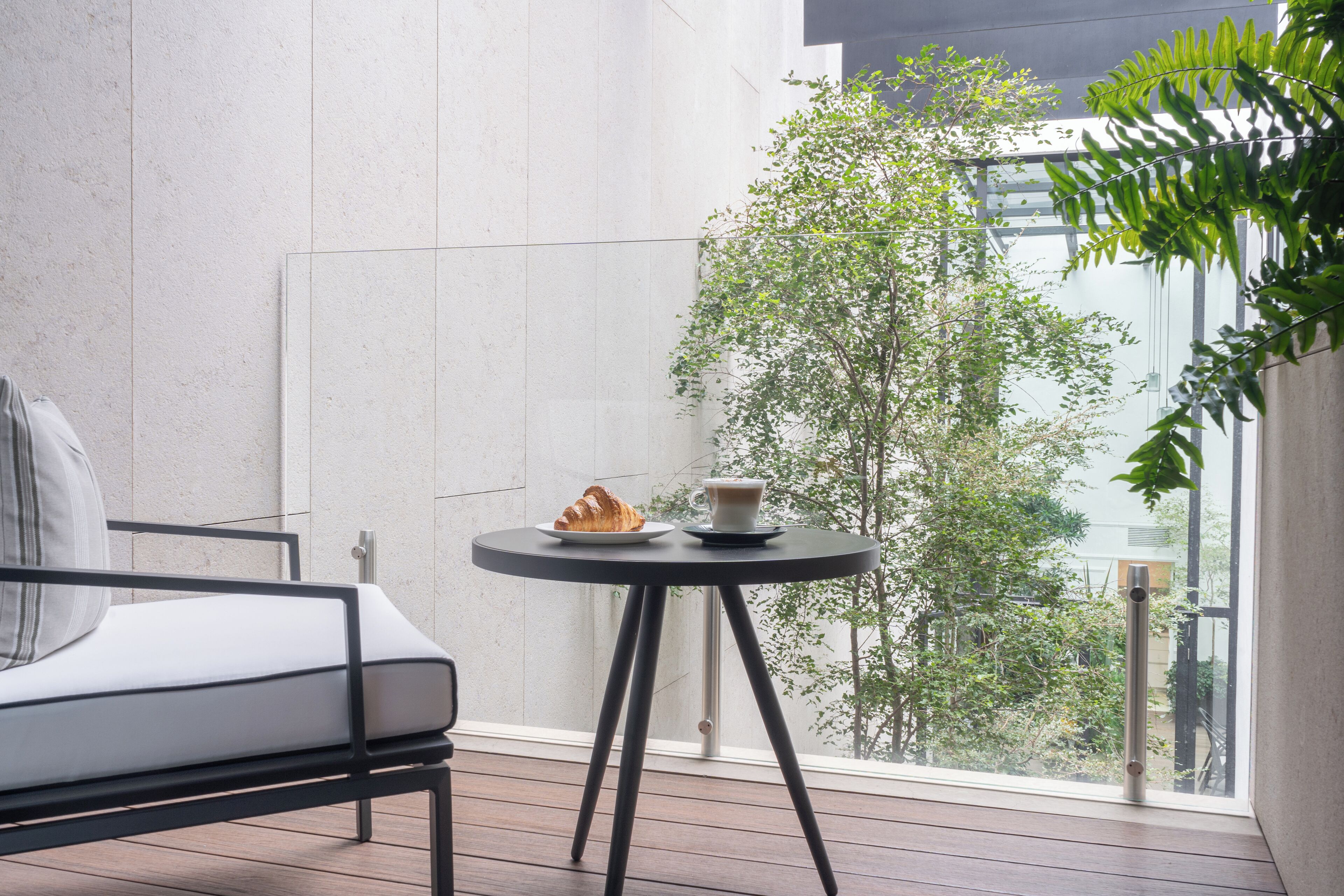 Un balcón moderno con vistas a árboles y edificios, una mesa redonda con un croissant y una taza, y una silla minimalista.