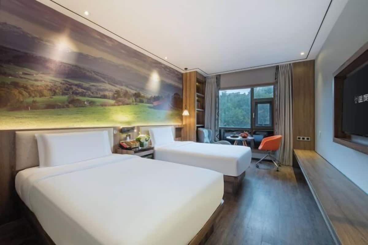 Habitación de hotel con dos camas, un escritorio, una silla, un ventanal grande y un cuadro en la pared.