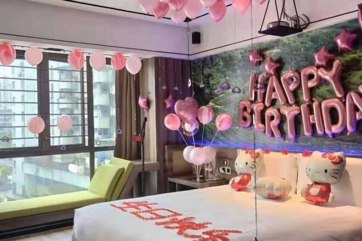 Una habitación con cama, ventana y pared decorada con globos de 'Feliz Cumpleaños' y un adorno de Hello Kitty.