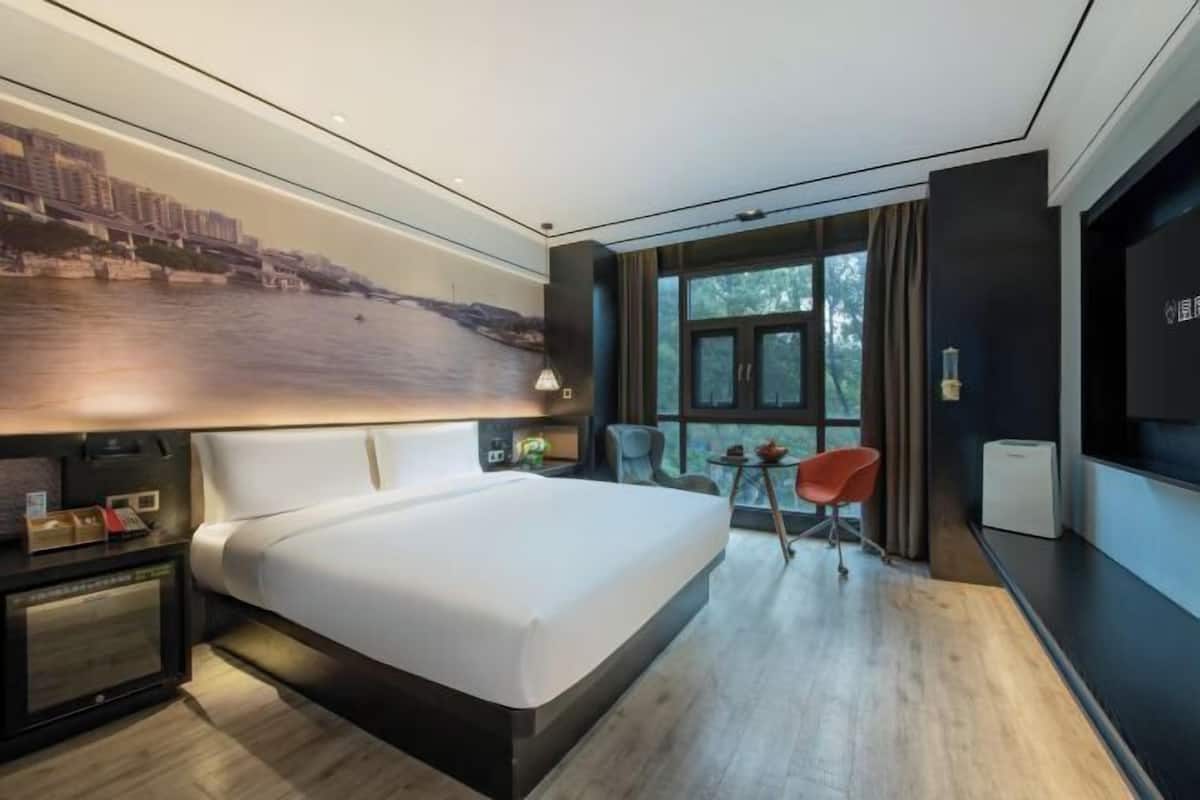 Una habitación de hotel moderna con una cama grande, un escritorio con una silla, un televisor y un ventanal con vistas a edificios y árboles.