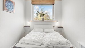 Een slaapkamer met een groot raam, een nachtkastje, een bedlampje, een bed met witte beddengoed en een schilderij aan de muur.