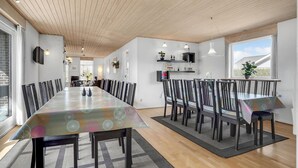 Een ruime eetruimte met een lange tafel en meerdere stoelen, houten plafond en grote ramen.