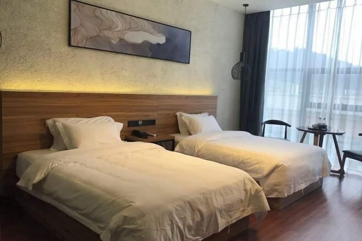 Hotellihuone, jossa on kaksi sänkyä, puinen pääty, taulu seinällä, pieni pöytä ja suuri ikkuna, josta avautuu näkymä kaupunkiin.