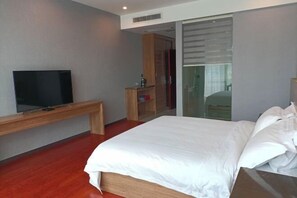 A modern hotel room with a large bed, a flat-screen TV on a wooden console table, a sliding glass door to a bathroom, and a small wooden cabinet. - Nanyuan Hotel (Jinsha)