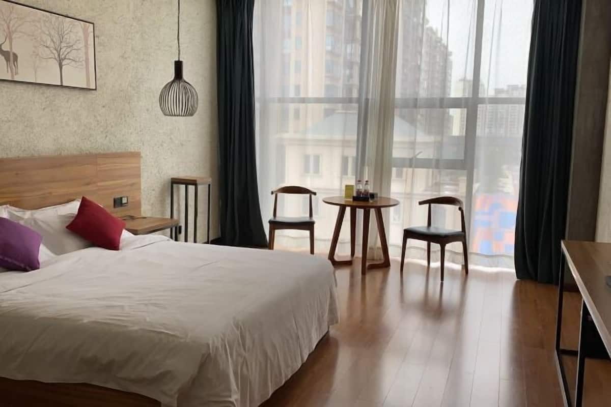 Hotellihuone, jossa on suuri sänky, puinen pääty, pieni pöytä tuoleineen ja näkymä rakennuksiin ohuiden verhojen läpi.
