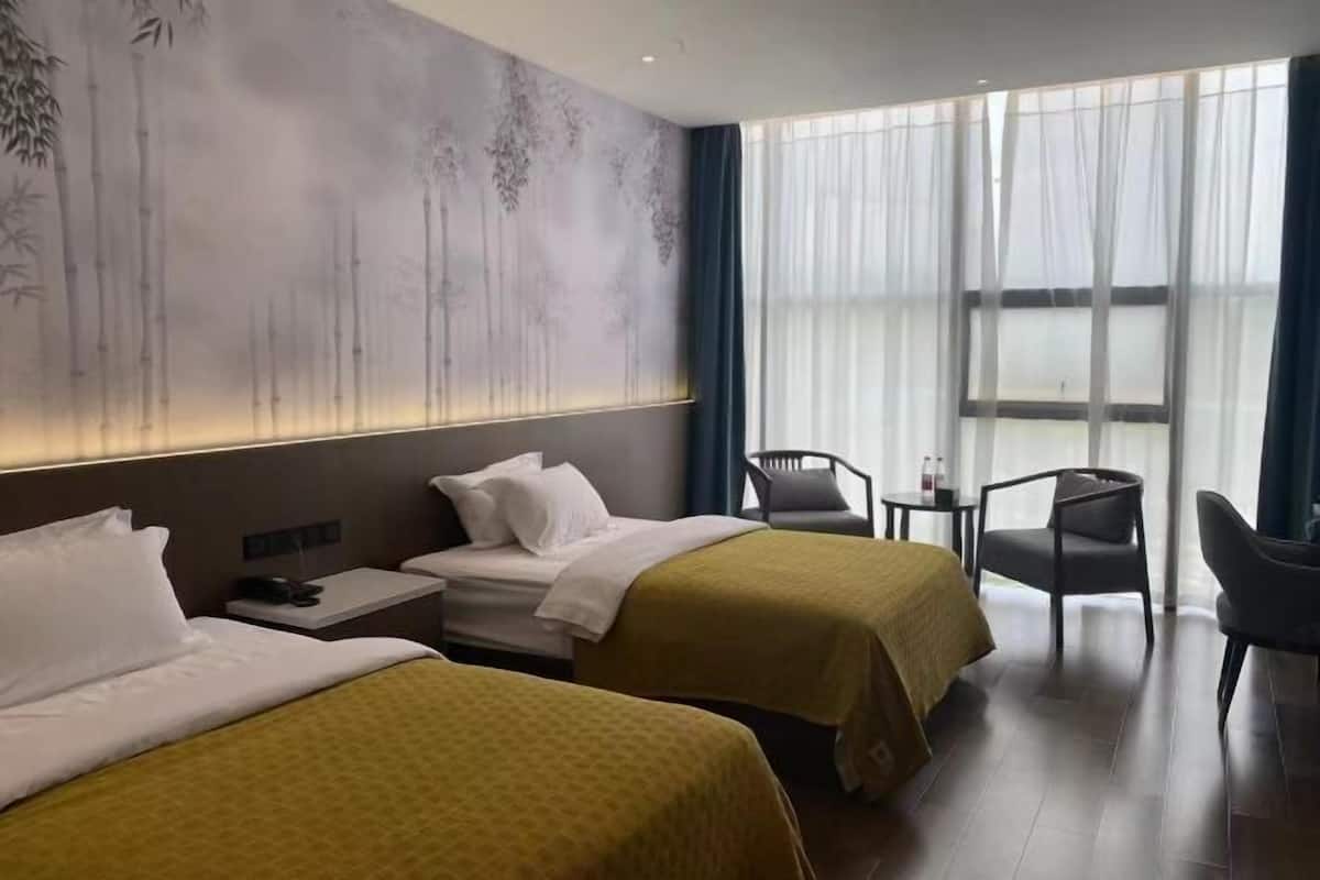 Hotellihuone, jossa on kaksi sänkyä, pieni pöytä, tuoli ja suuri ikkuna, jossa on ohuet verhot.