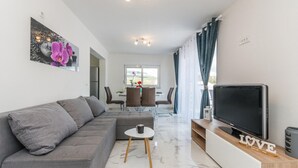 Una sala de estar moderna con un sofá gris, un televisor sobre un mueble de madera y un comedor al fondo.