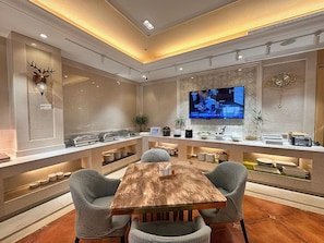 A modern lounge area with a wooden table, cushioned chairs, a flat-screen TV, and a decorative deer head on the wall.
