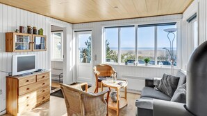 A cozy living room with a wooden ceiling, a large window overlooking a beach, a grey sofa, wicker chairs, and a wooden cabinet with glass doors. - 2 bedroom beautiful home in Rømø (Romo)