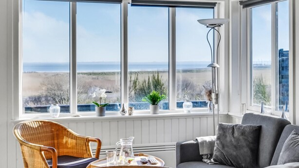 A room with a large bay window offering a view of the beach and ocean. There is a wicker chair, a grey sofa, and a table with a tray of items. - 2 bedroom beautiful home in Rømø (Romo)