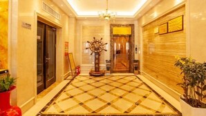 Een hotellobby met een zwart-wit geruite vloer, een kroonluchter, een lift en een muur met ingelijste certificaten.